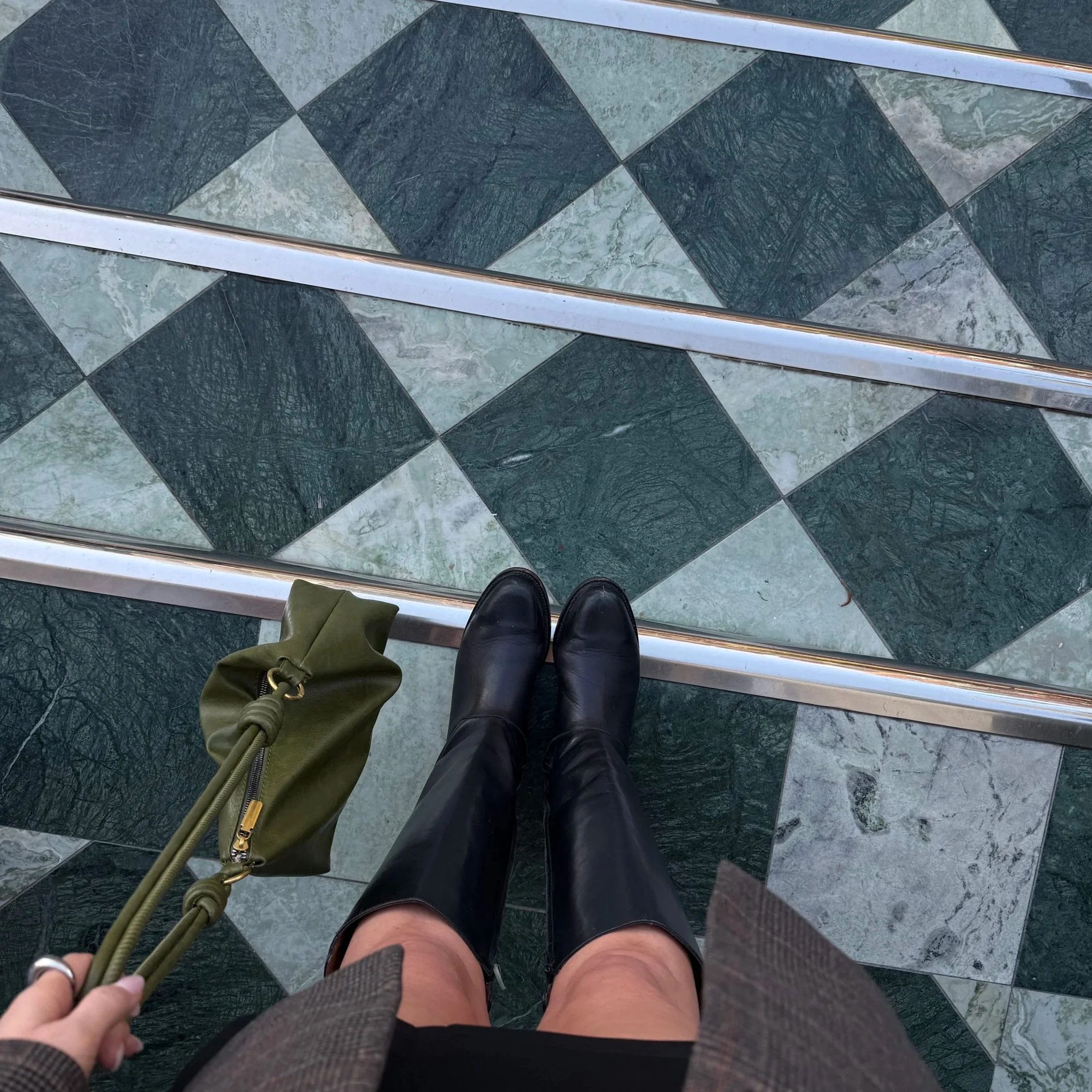 View of a person's legs in black leather boots standing on a tile floor with green and gray marble pattern. The person is holding a green handbag with gold zipper and clasp. The photo is taken from a top-down perspective.