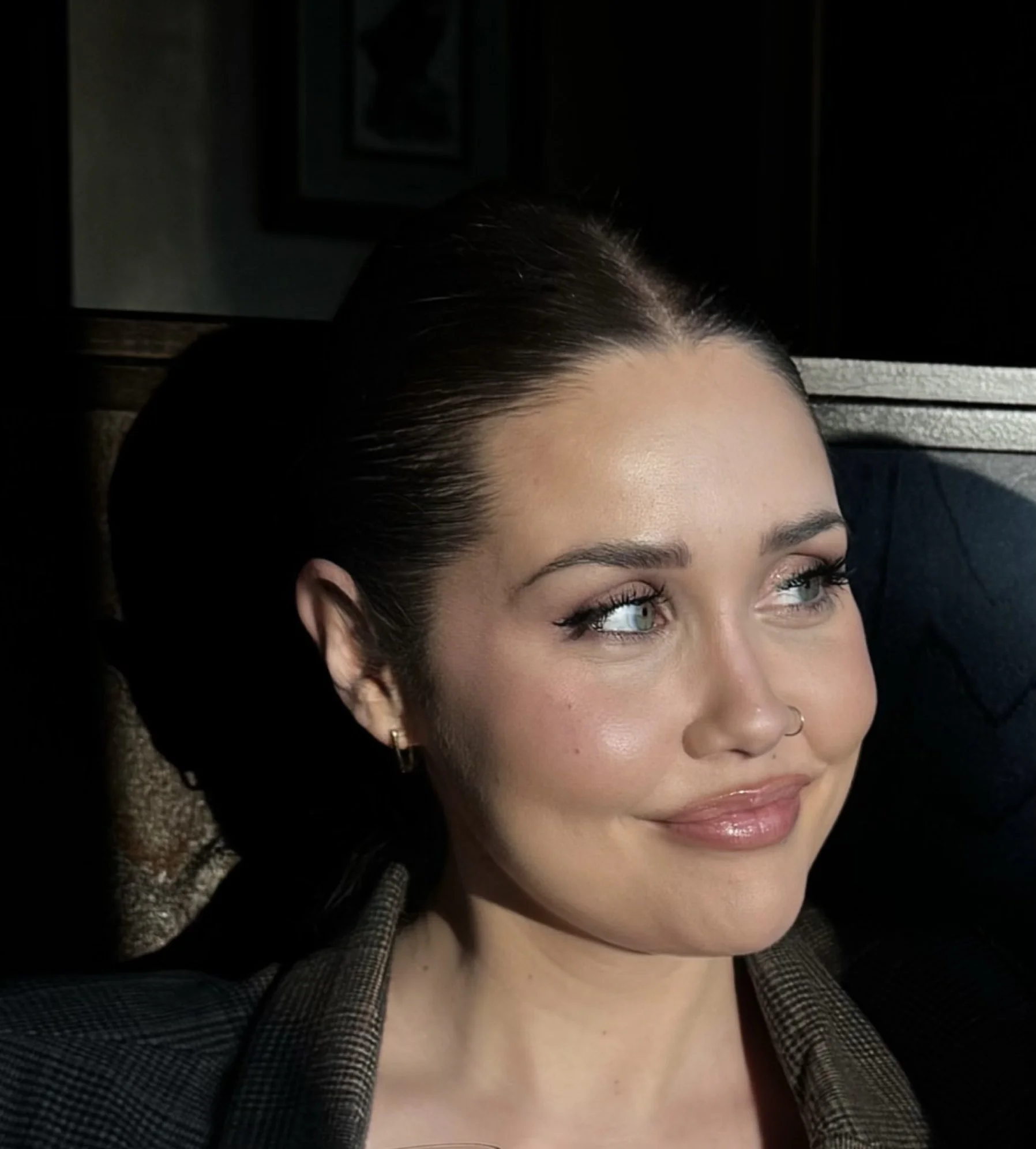 Close-up of a woman with dark hair, light skin, and blue eyes, smiling softly and looking to the side, with sunlight illuminating her face.