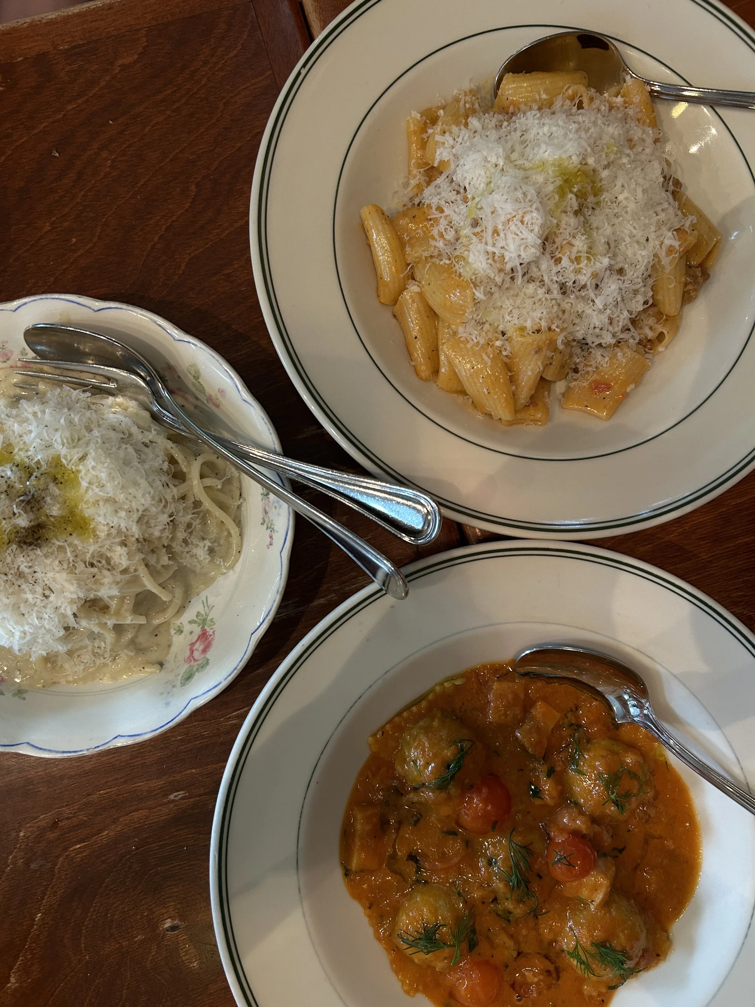 Three dishes of pasta and a vegetable curry served on white plates with silver utensils, on a wooden table.
