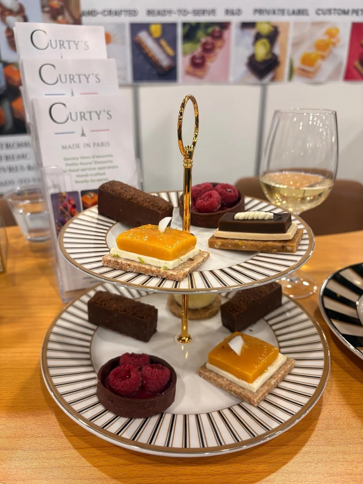 Two tier stand of French petits fours with mango tartlets, raspberry chocolate tarts, chocolate cake bars and caramel domes beside a glass of white wine