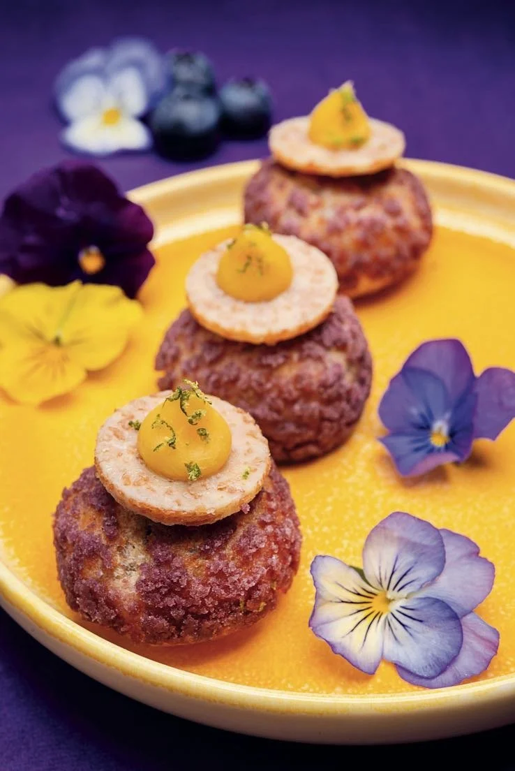 Round French mini puffs topped with orange and edible pansies on a yellow plate, photographed from above