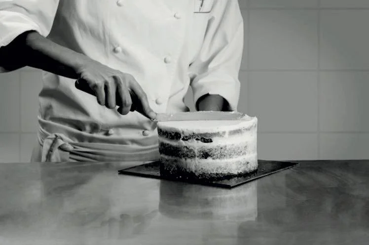 Black and white photo of a pastry chef in whites finishing a layered cake with a spatula on a stainless steel counter