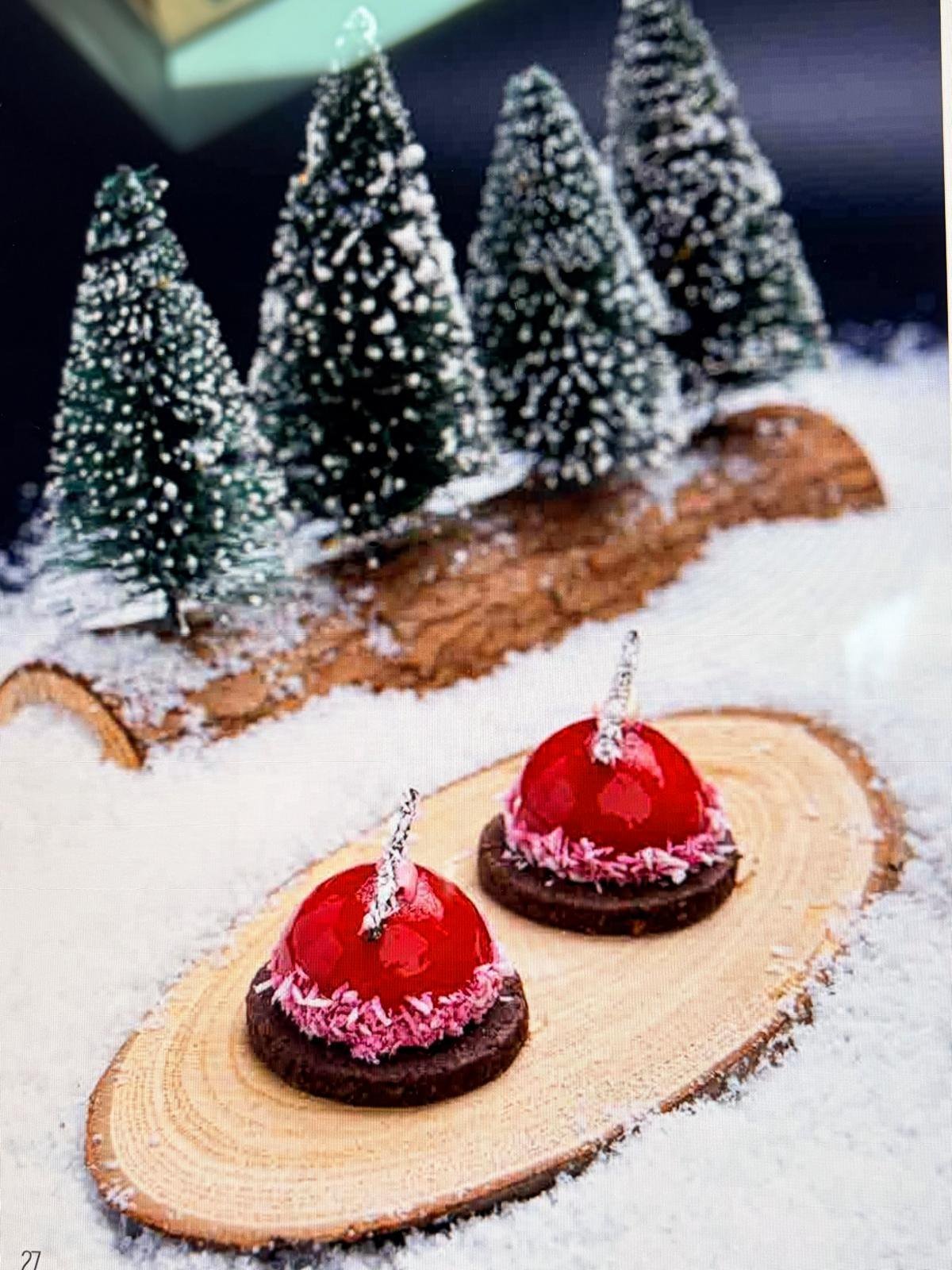 Two red glazed raspberry chocolate coconut domes on a chocolate biscuit base with pink coconut at the edge, served on a wood slice with miniature snowy trees