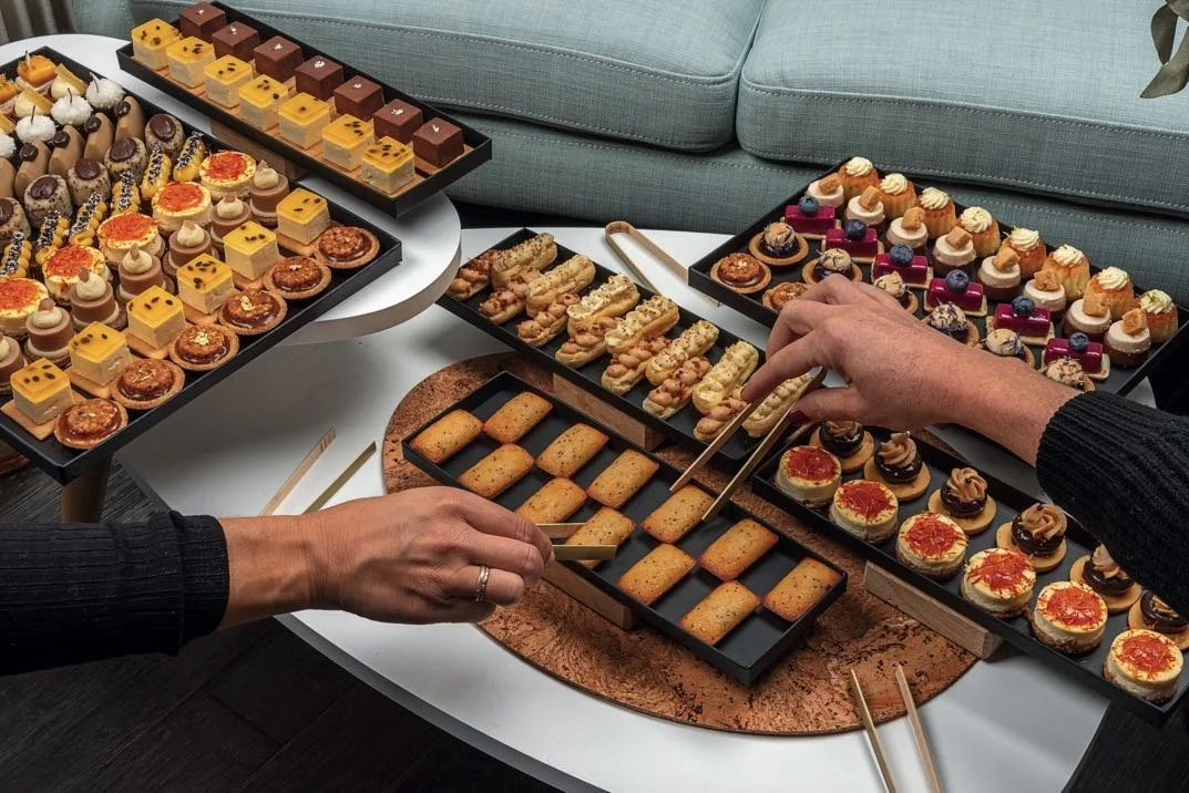 Guests picking French petits fours and financiers with tongs from black serving trays on a lounge coffee table