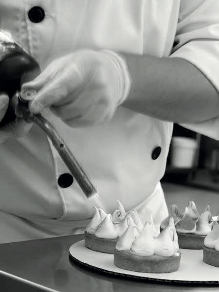 Black and white photo of a pastry chef in gloves torching meringue topped tartlets on a cake board