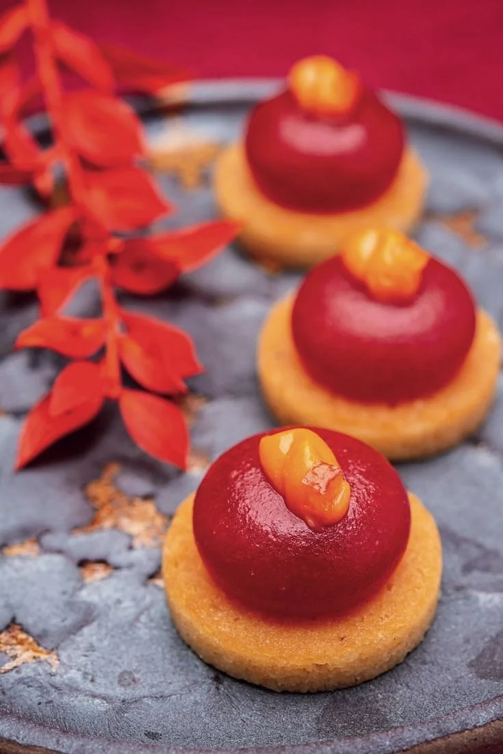 Three glossy red glazed petits fours on round biscuit bases, topped with an orange garnish, on a dark plate with red leaves