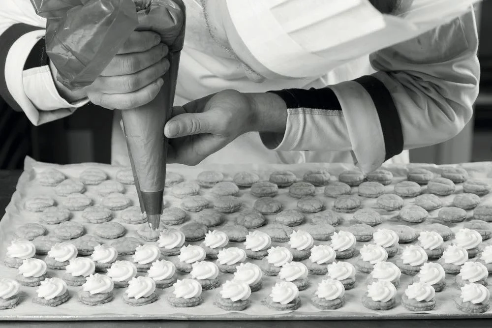 Black and white photo of a pastry chef piping cream onto rows of small pastry shells on a baking tray