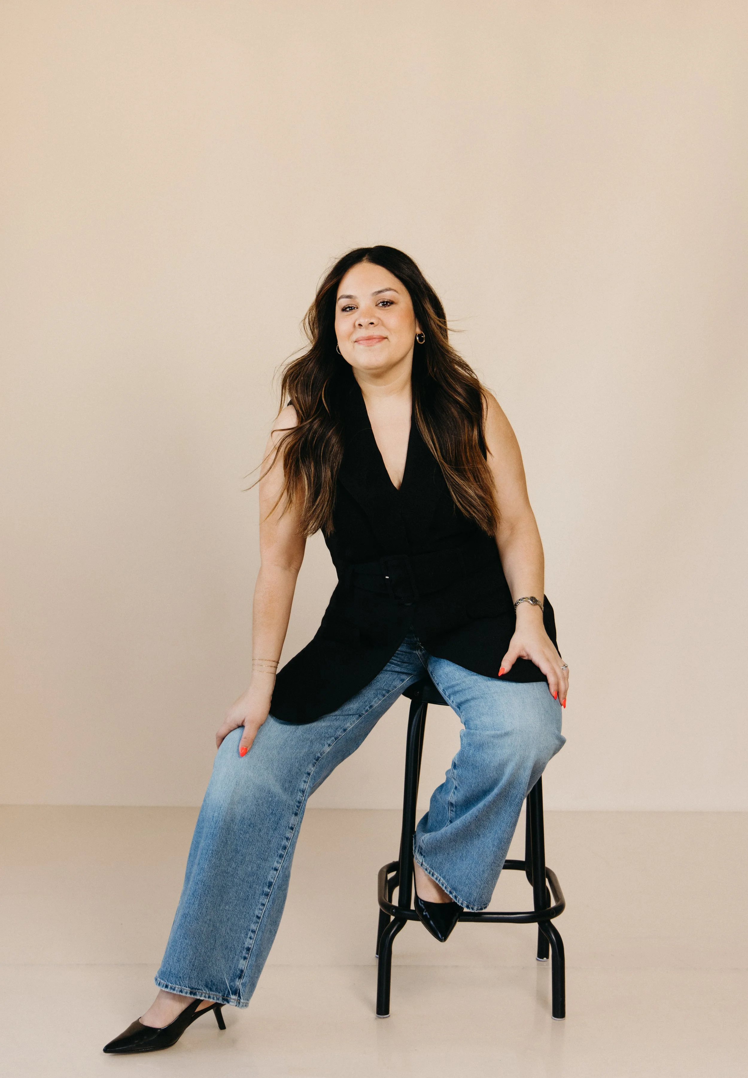Aimee Bruno sitting on a black stool, wearing a sleeveless black top, wide-leg jeans, and black high heels, against a beige background.
