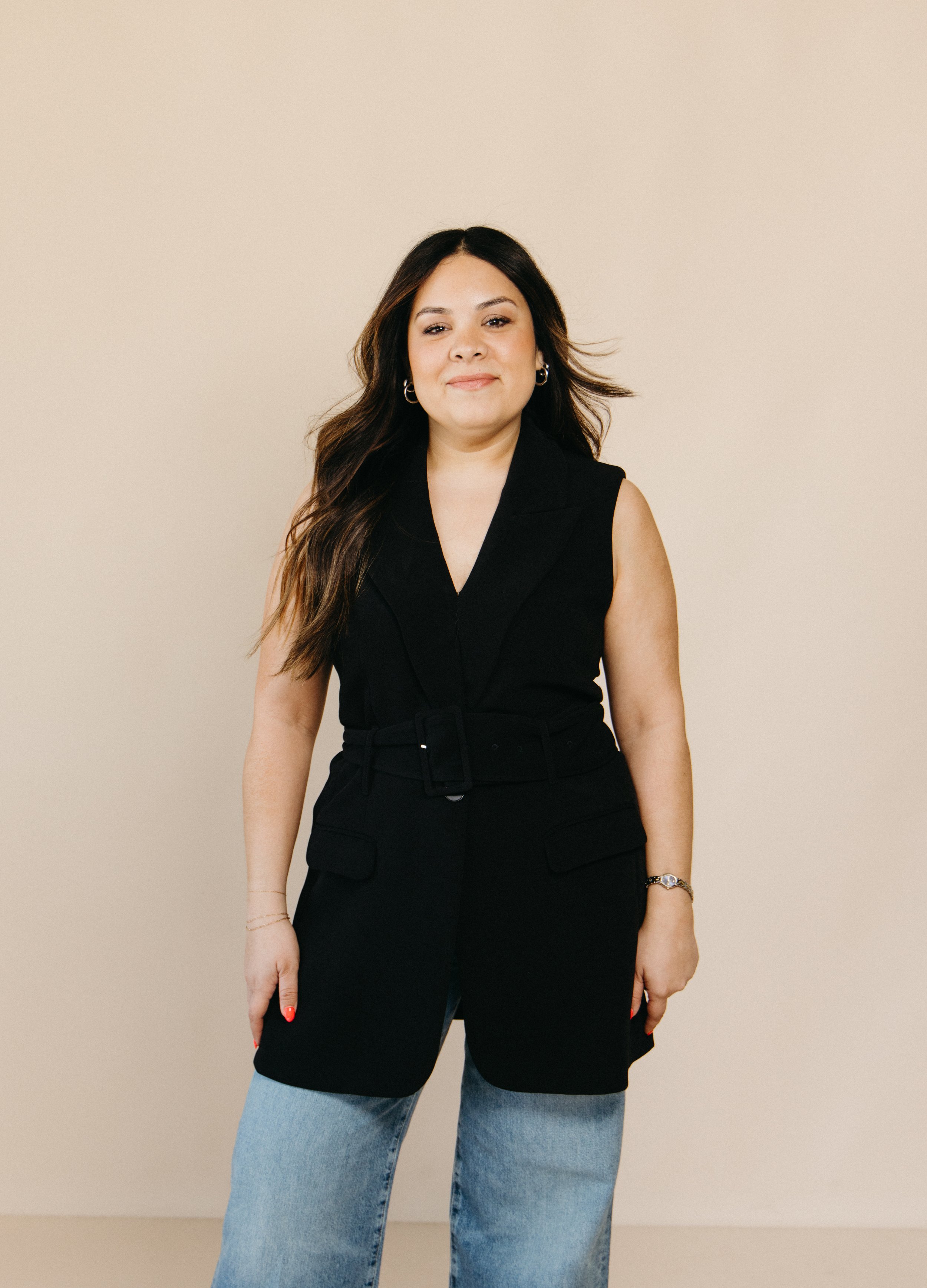 Aimee Bruno wearing a black sleeveless blazer top and light blue jeans standing against a plain beige wall.