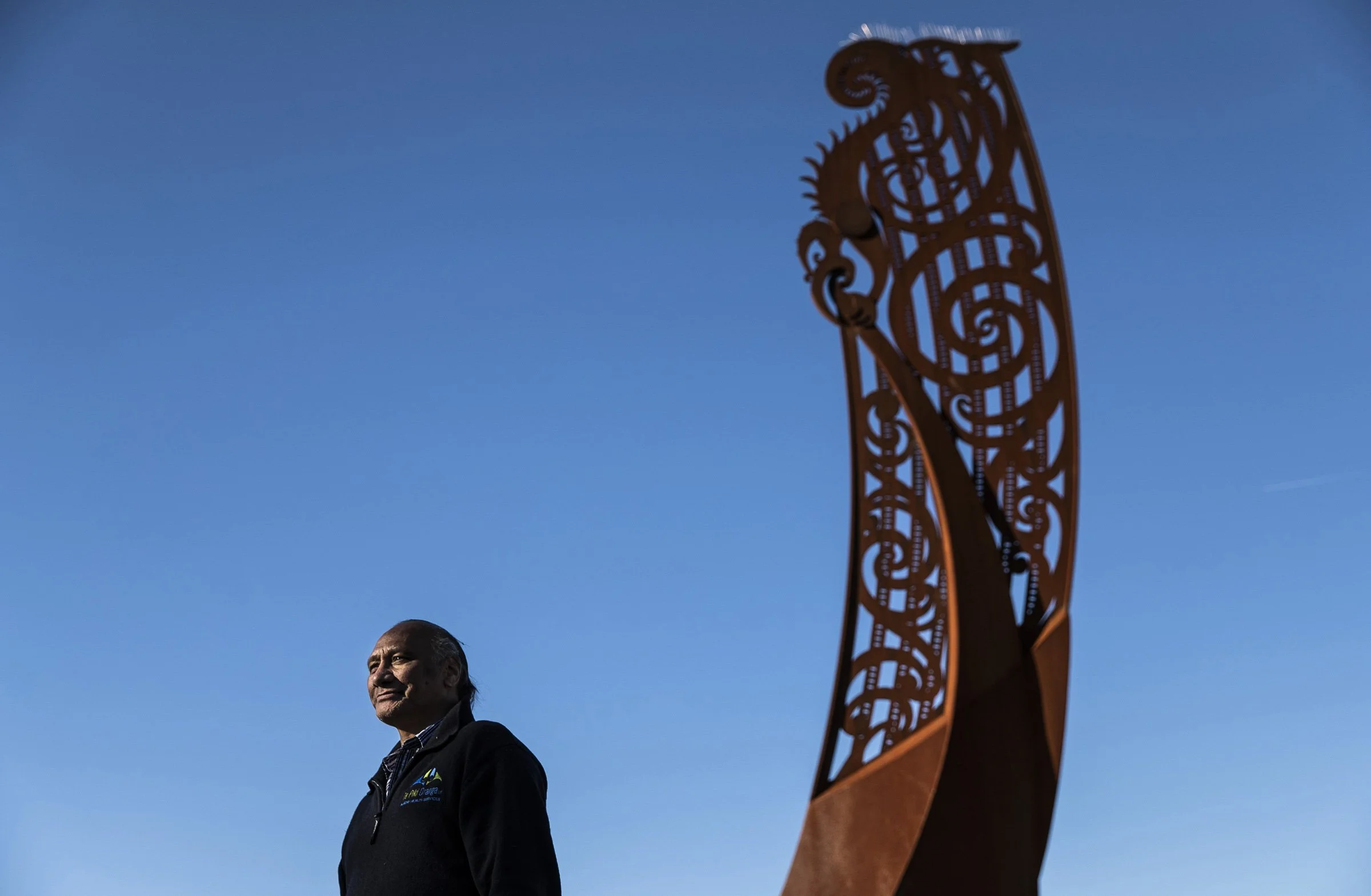 A man smiling outdoors, standing near a carved wooden sculpture in the background.