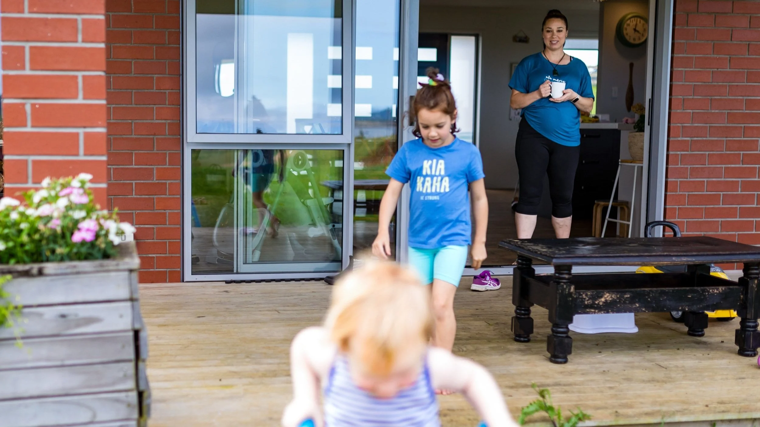 Young girl in blue shirt and shorts walking on porch, holding a toy, with a woman in a blue shirt and black pants holding a mug in the background, standing inside a house near open sliding door, with another child in the foreground and outdoor plants visible.