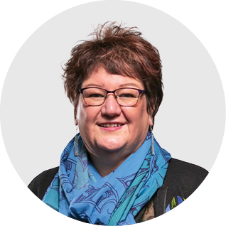 A woman with short curly brown hair, glasses, and a blue patterned scarf smiling against a plain background.