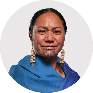 Portrait of a person with dark hair and facial tattoo, wearing earrings and a blue garment, against a white background.