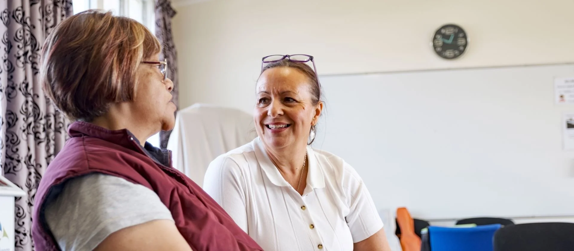 Two women having a conversation in a bright room, one with glasses and a white shirt, the other with short hair and a maroon vest, with a whiteboard and a clock in the background.