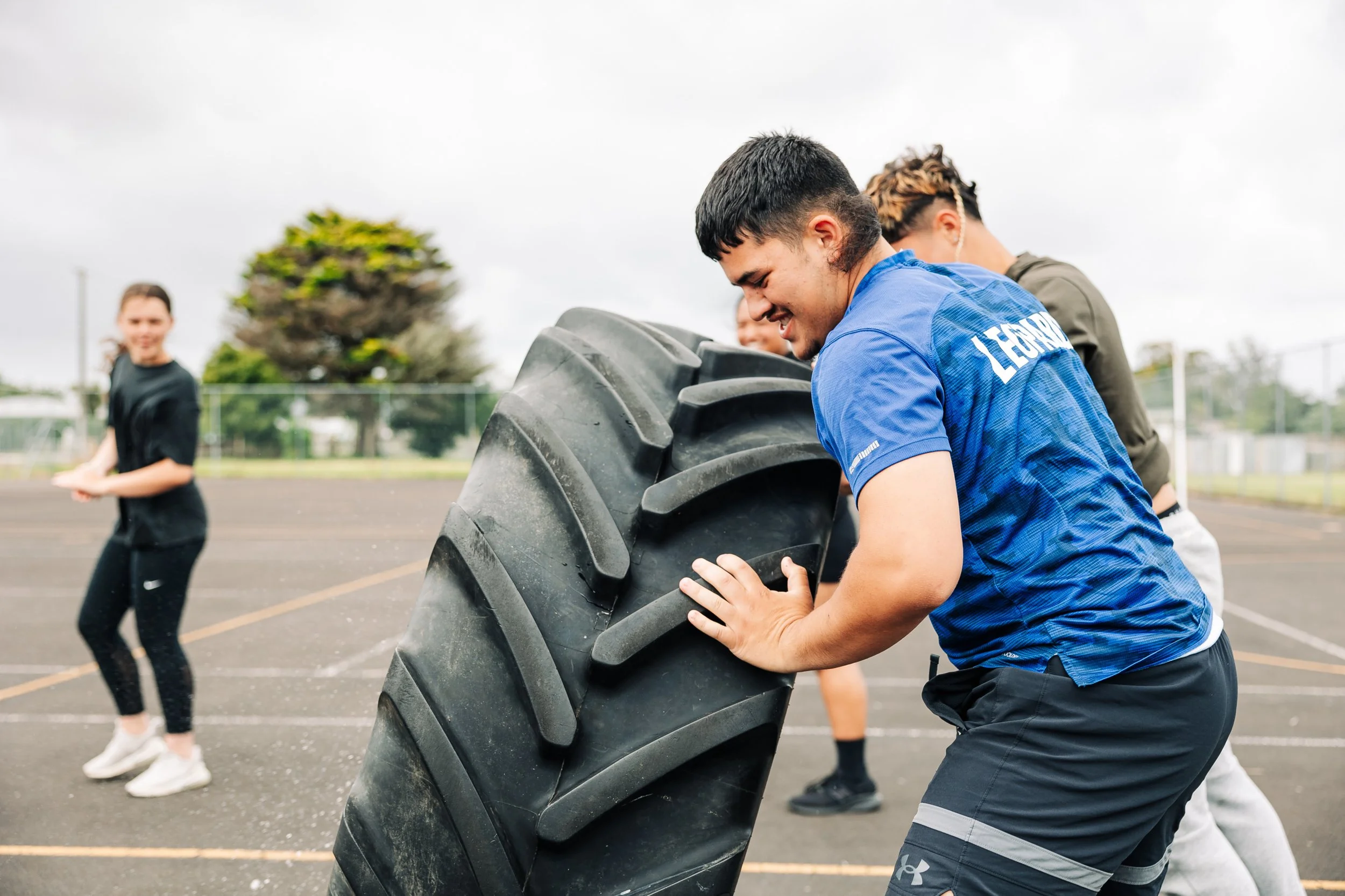 Young man in blue sports shirt lifting a large tire during outdoor workout, with other people in the background