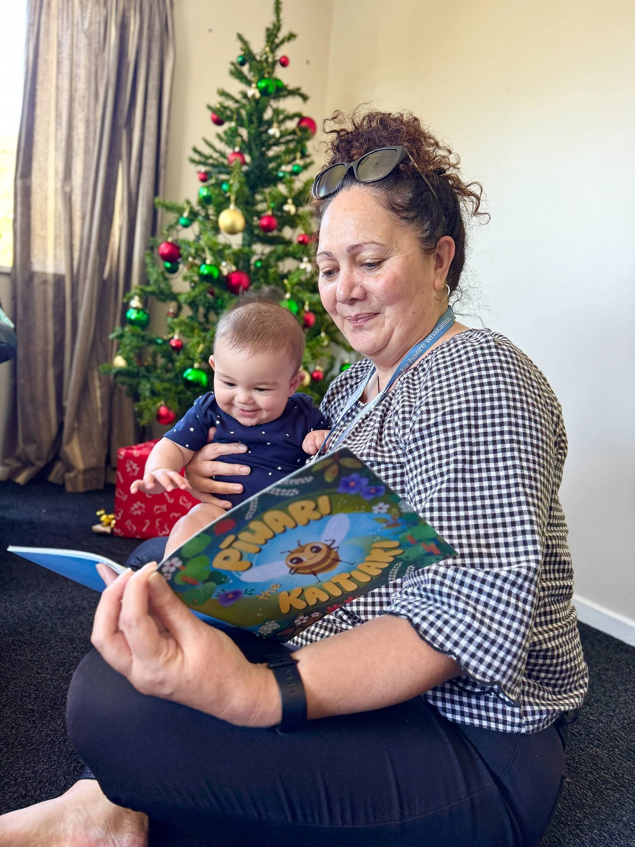 A woman with sunglasses on her head and curly hair is sitting on the floor, holding a smiling baby in her lap. They are reading a colorful children's book titled 'Pinari The Kauri' with a gnome and other illustrations on the cover. Behind them, there is a decorated Christmas tree with red, green, and gold ornaments, and a wrapped gift at the base.