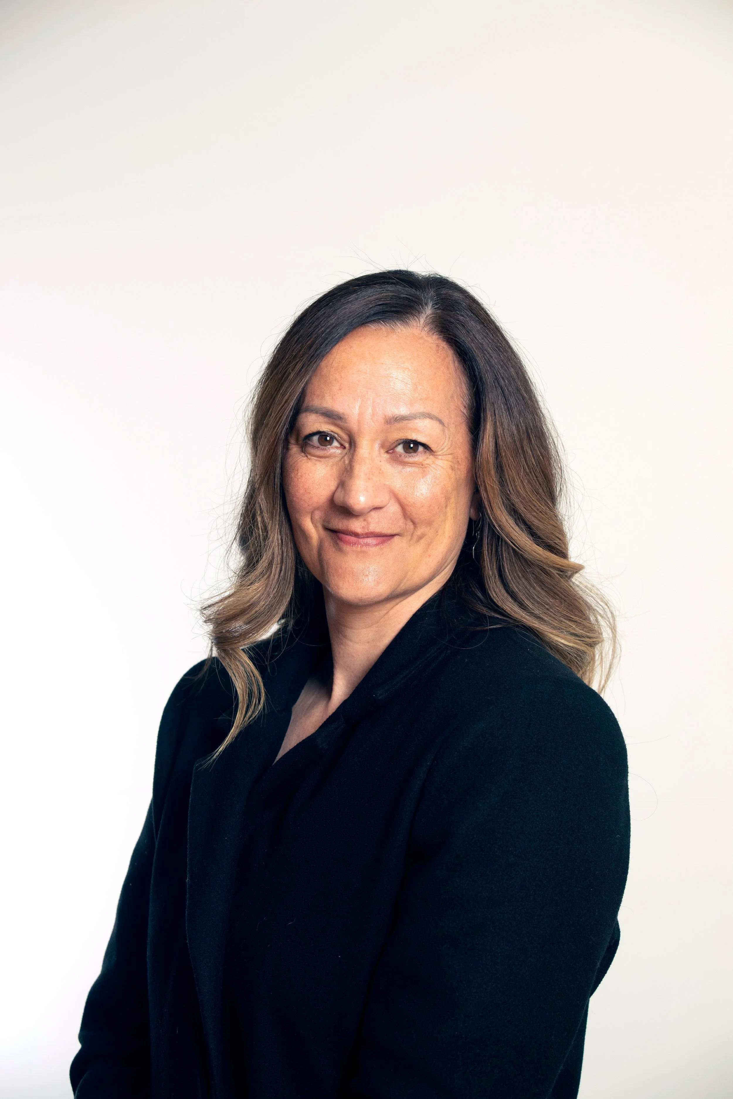 A woman with shoulder-length brown hair styled in loose waves, wearing a black blazer, is smiling softly and looking at the camera against a plain light background.