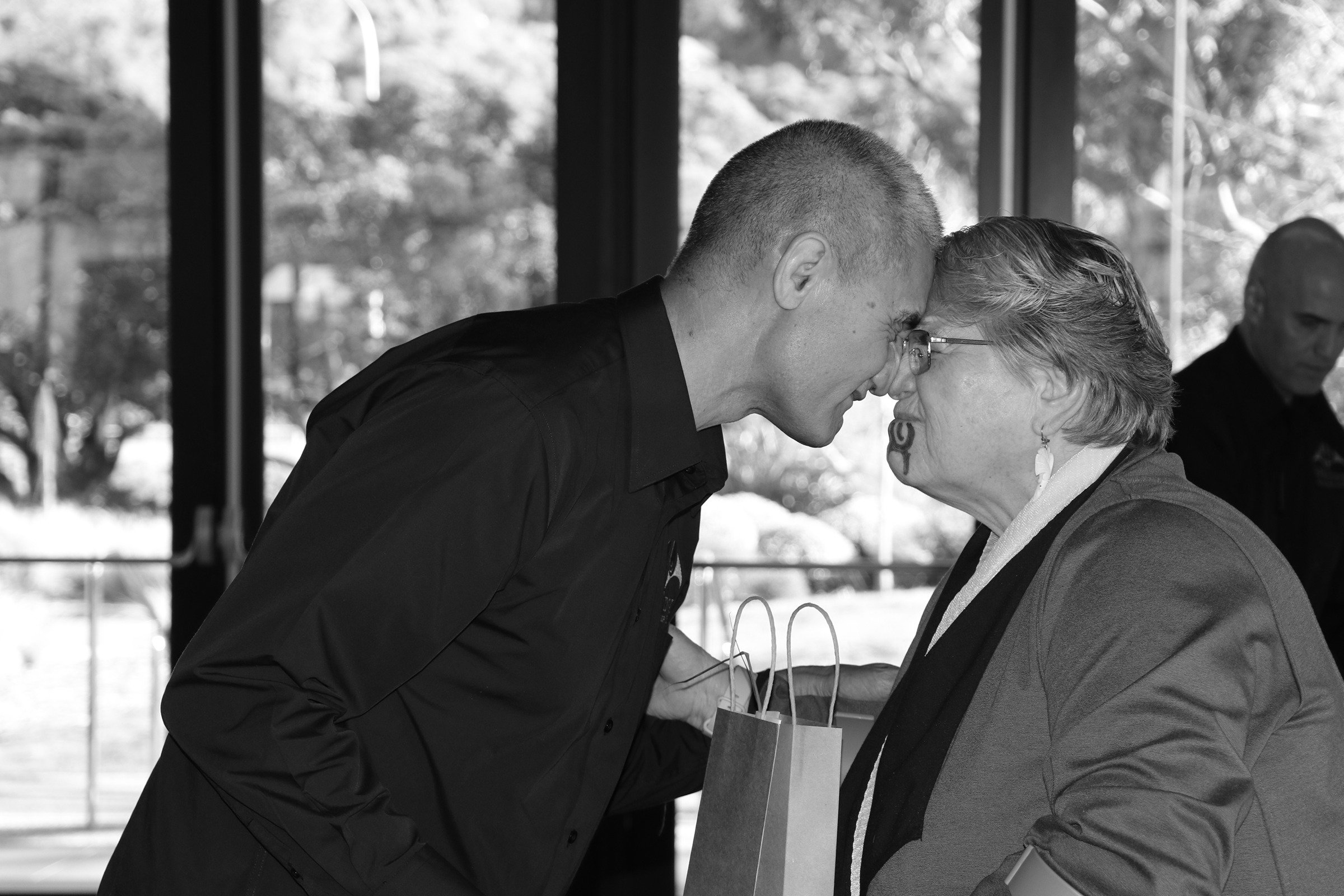 A young man and an older woman are touching foreheads and smiling at each other indoors, with trees visible outside through large windows.