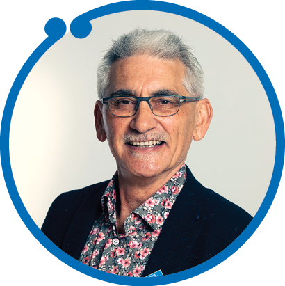 A smiling elderly man with glasses, gray hair, wearing a dark blazer and a colorful floral shirt, against a light background.