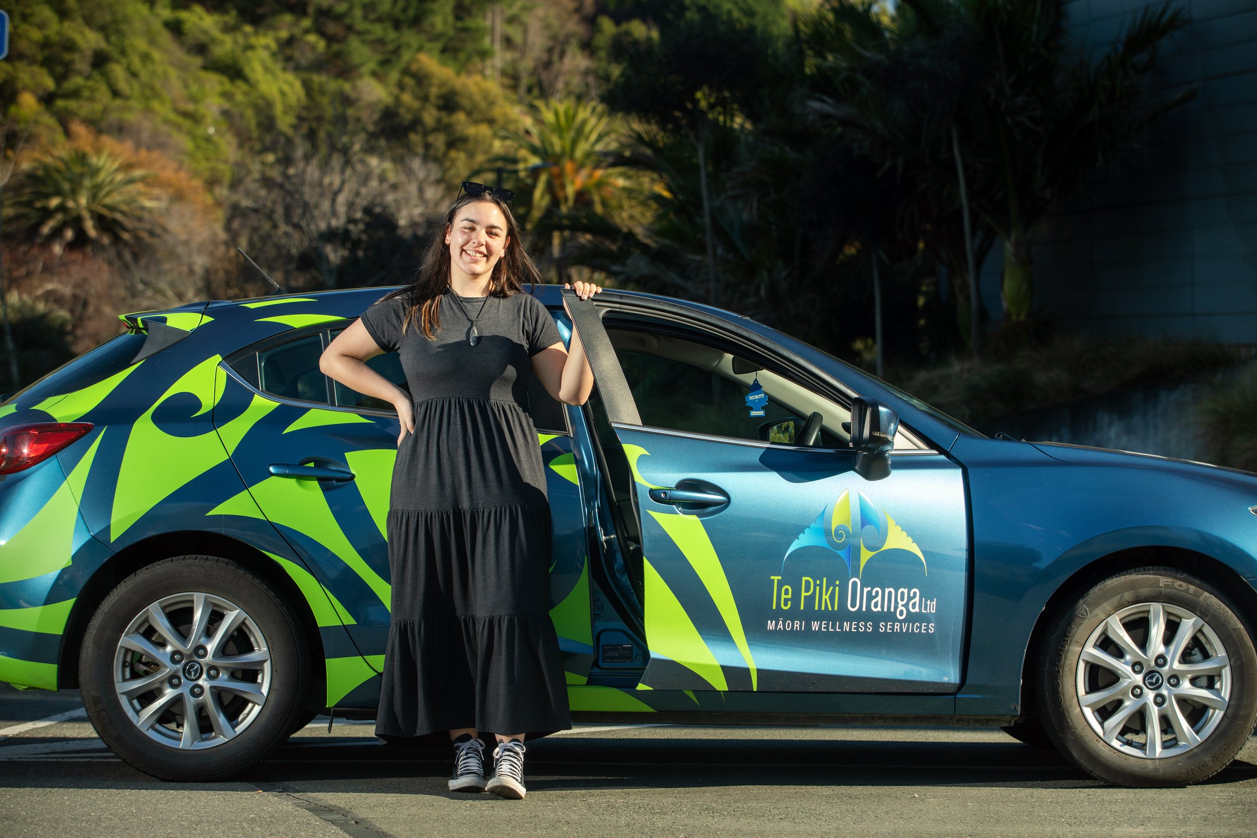 A young woman in a black dress and sneakers standing next to a colorful car with Maori wellness branding, outdoors on a sunny day.