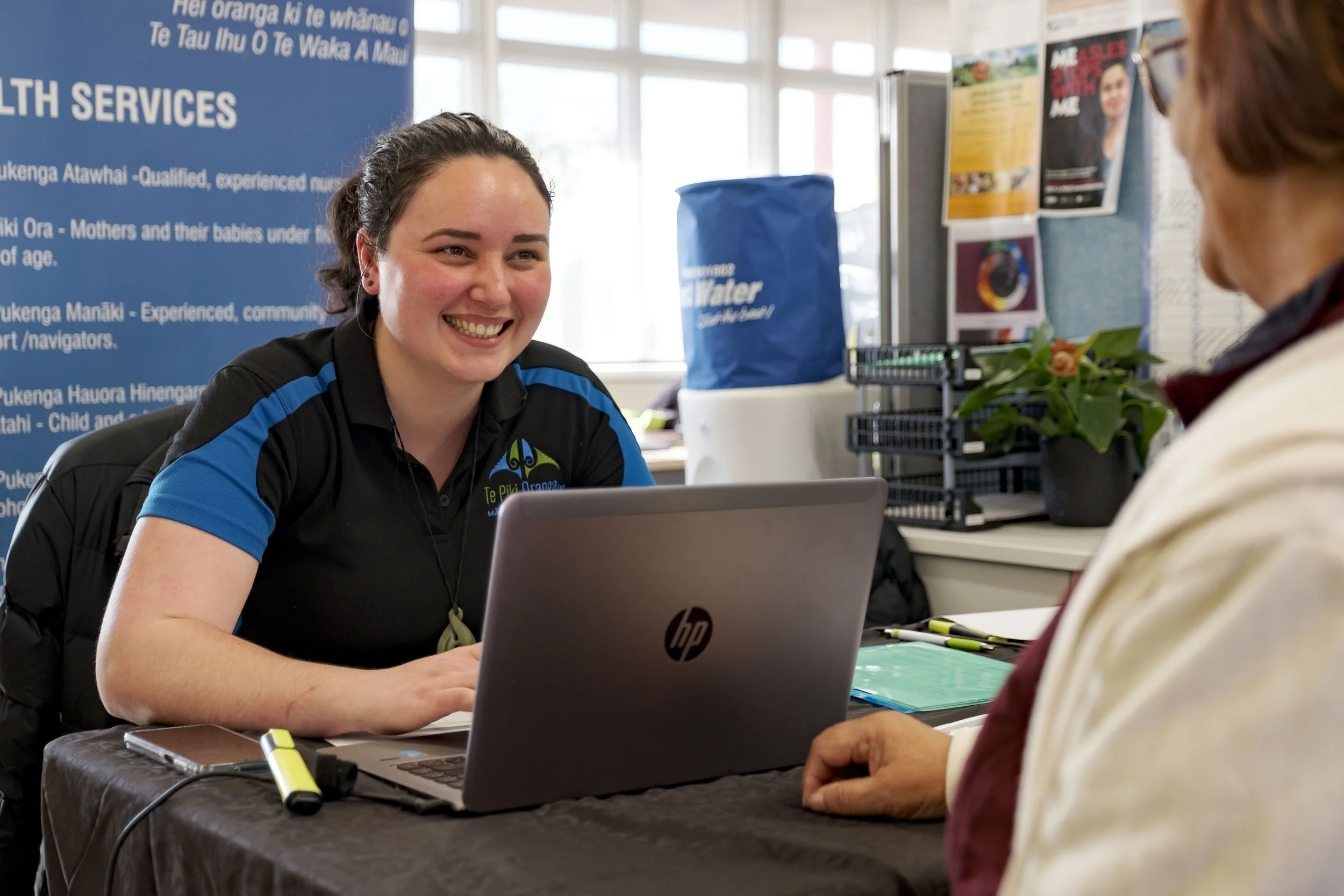 A woman with dark hair in a ponytail smiling while sitting at a desk with a laptop, talking to an older woman. There is a large blue height chart with text behind her and various informational posters on the wall.