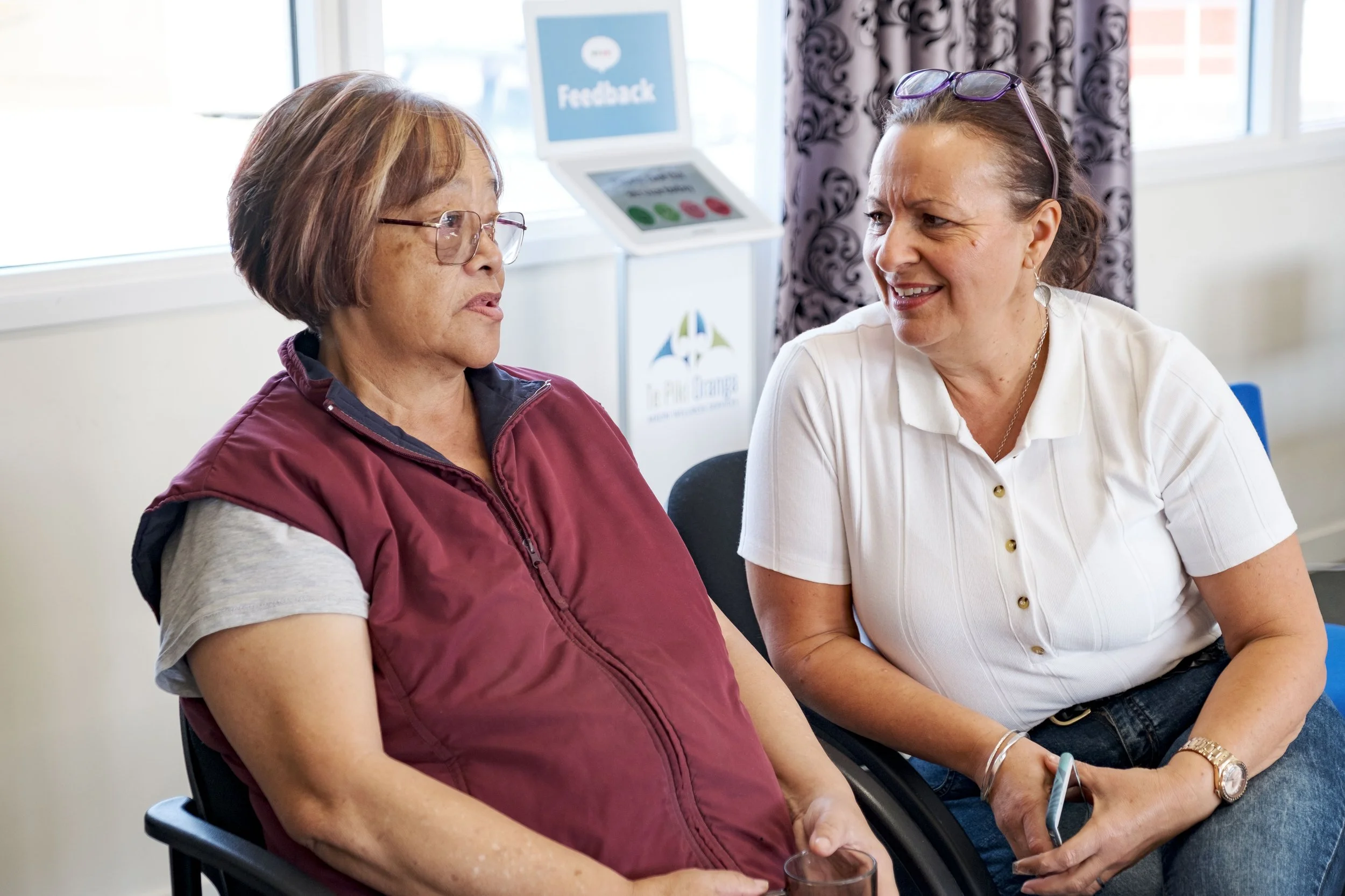 Two women having a conversation in an office or waiting room, with feedback signage in the background.