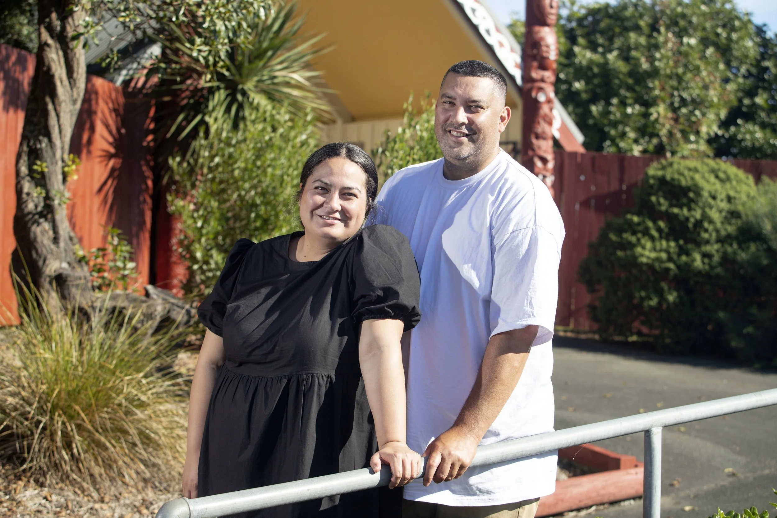 A smiling couple standing outside in front of a red fence and lush green bushes, with a decorative building in the background.