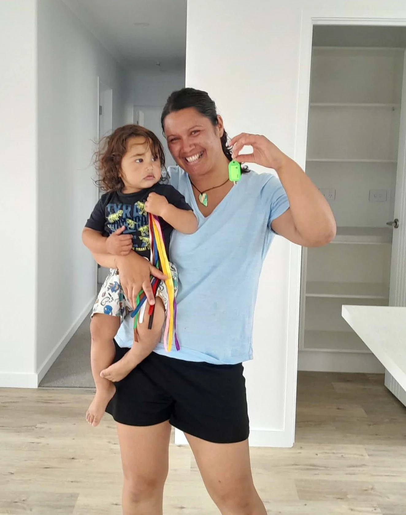 A woman is smiling and holding a young child, who is holding colorful ribbon ties, inside a house.