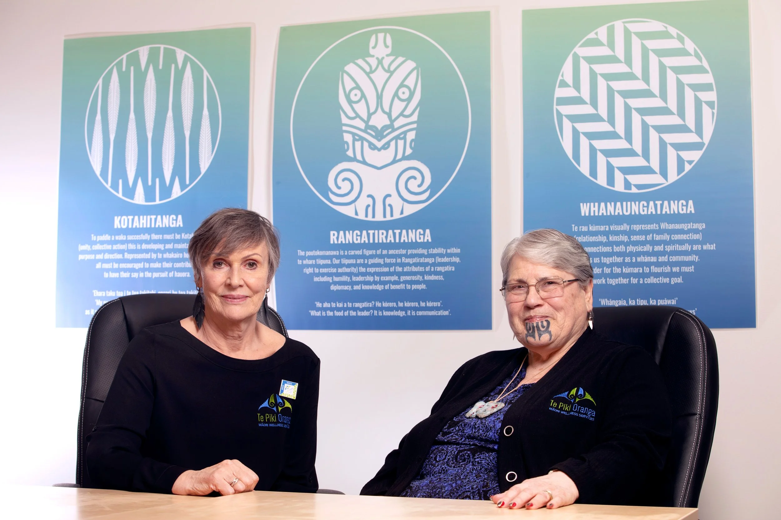 Two women sitting at a table in front of three blue posters with Māori symbols and text. The woman on the left has short gray hair and is wearing a black shirt with a logo. The woman on the right has gray hair, glasses, tattoos on her face, and is also wearing a black shirt with a logo.