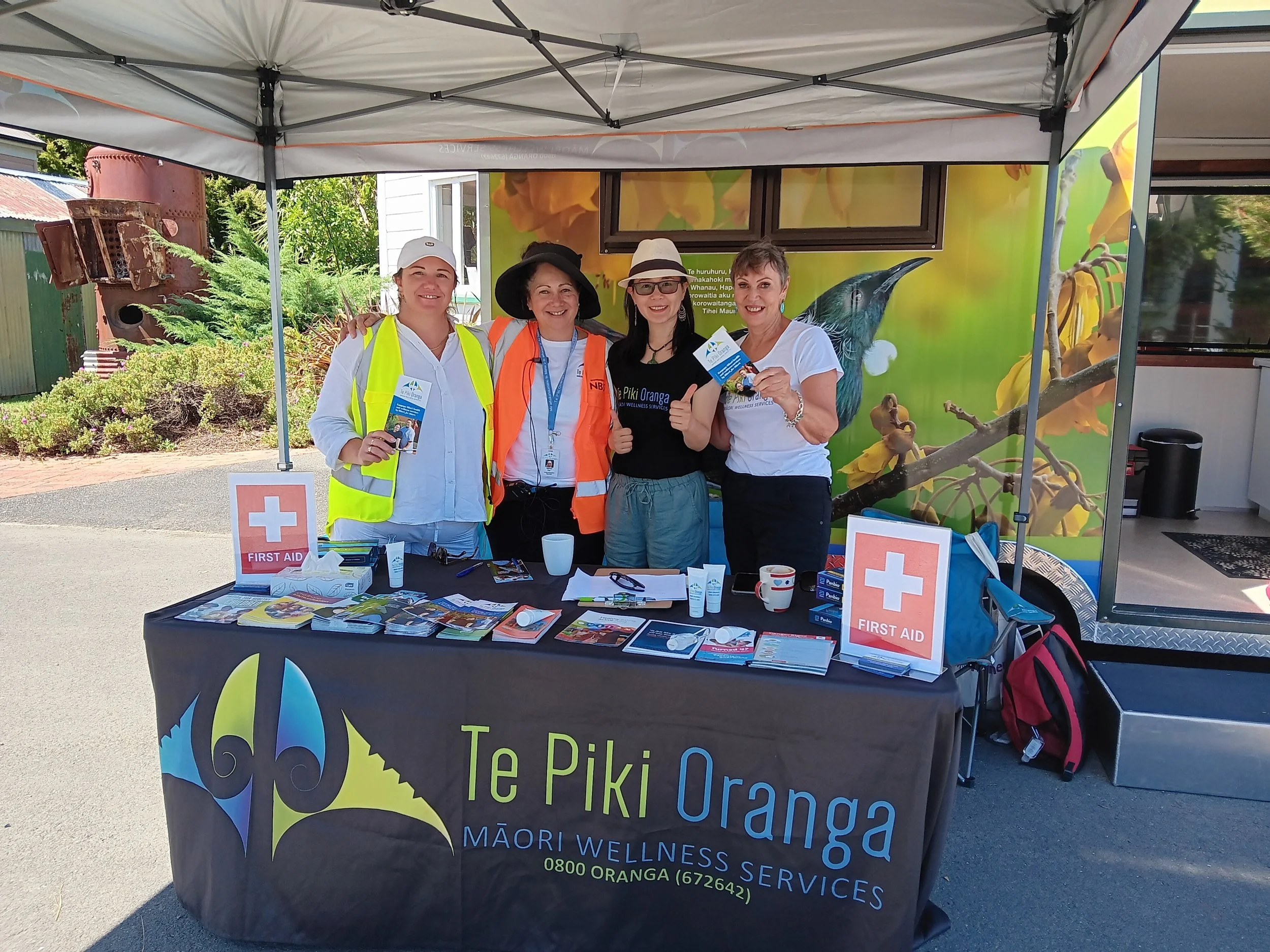 Four women standing behind a table at an outdoor booth. They are smiling and holding pamphlets. The table has First Aid signs and various pamphlets and supplies. The booth is under a canopy, with a colorful backdrop featuring birds and flowers. The women appear to be promoting Maori wellness services.