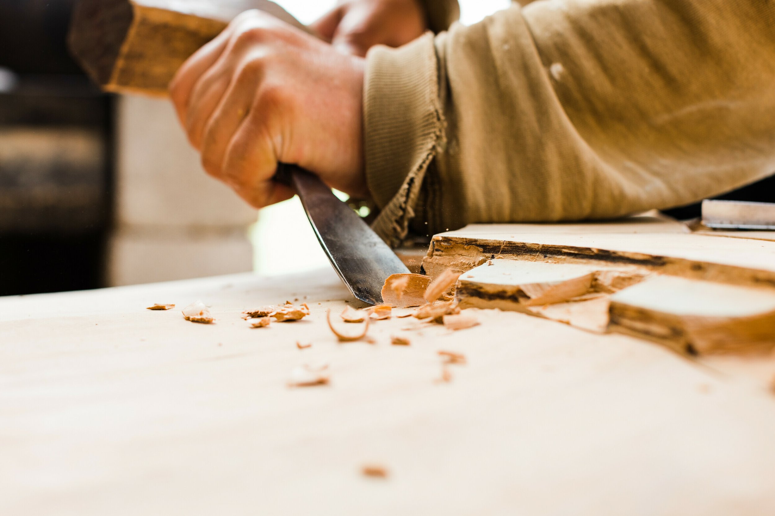 Close-up of a person's hand using a knife to carve wood on a workbench with wood shavings.