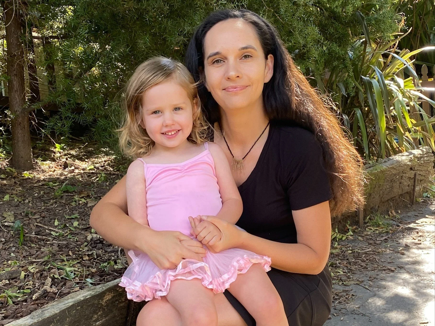 A woman with long black hair and a young girl with curly blonde hair sitting together outdoors surrounded by green plants. The woman is wearing a black shirt and a necklace, while the girl is in a pink dress with ruffles.