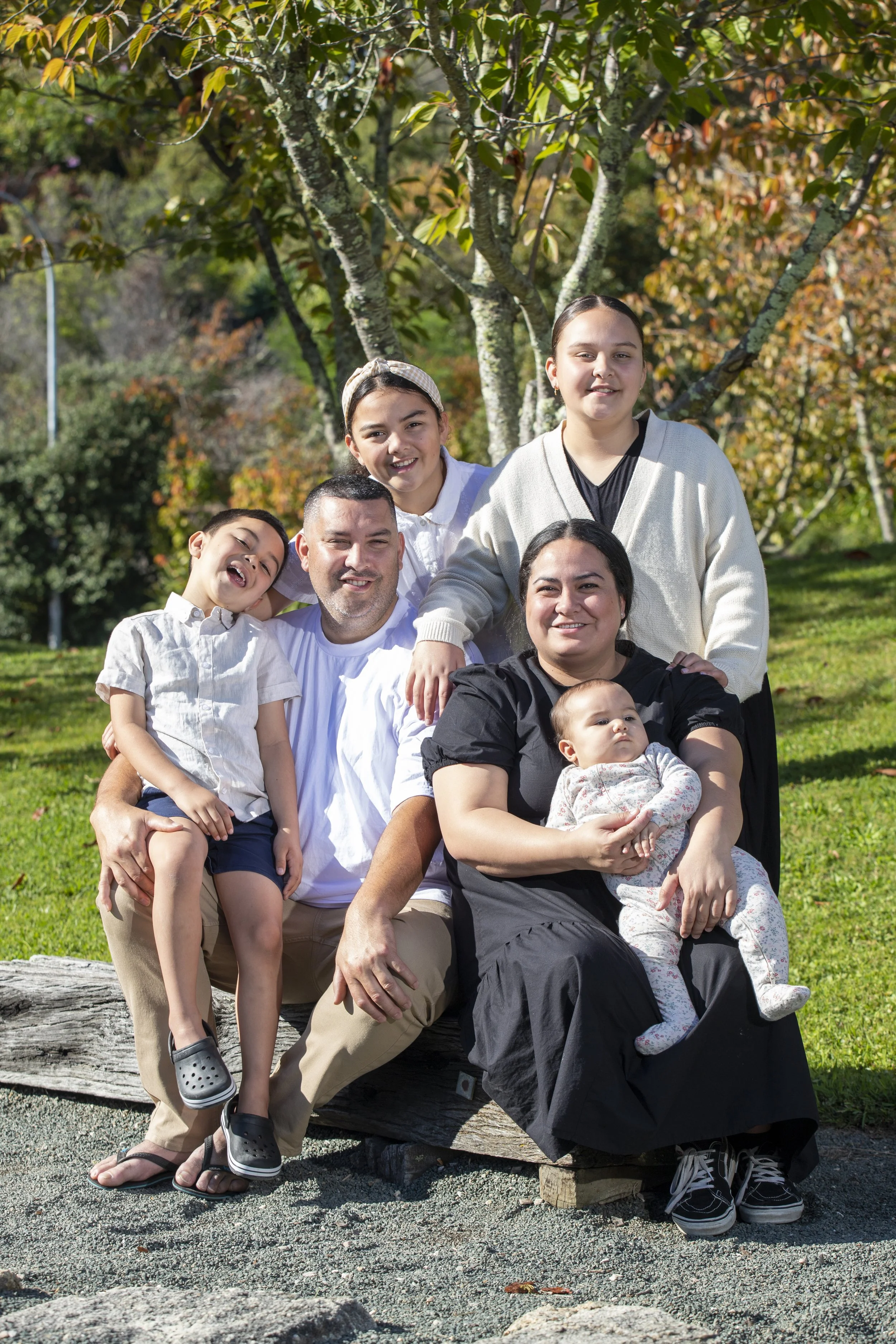 A family of seven posing outdoors on a sunny day, sitting on a wooden log with trees and greenery in the background.