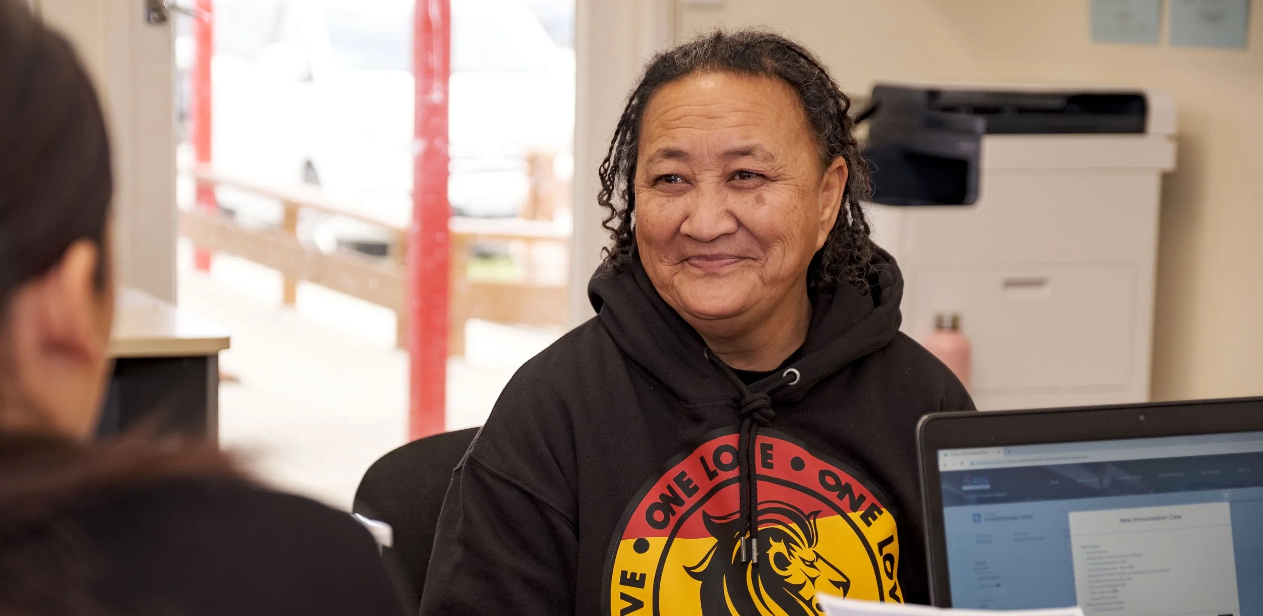 A man with curly hair and a black hoodie smiling at a woman in a black jacket, seated at a table in an indoor setting with a computer in front of her.