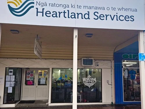 Exterior of a Heartland Services building with large sign displaying its name. The building has a glass front with posters and offers various services, located in a shopping area with neighboring stores visible.