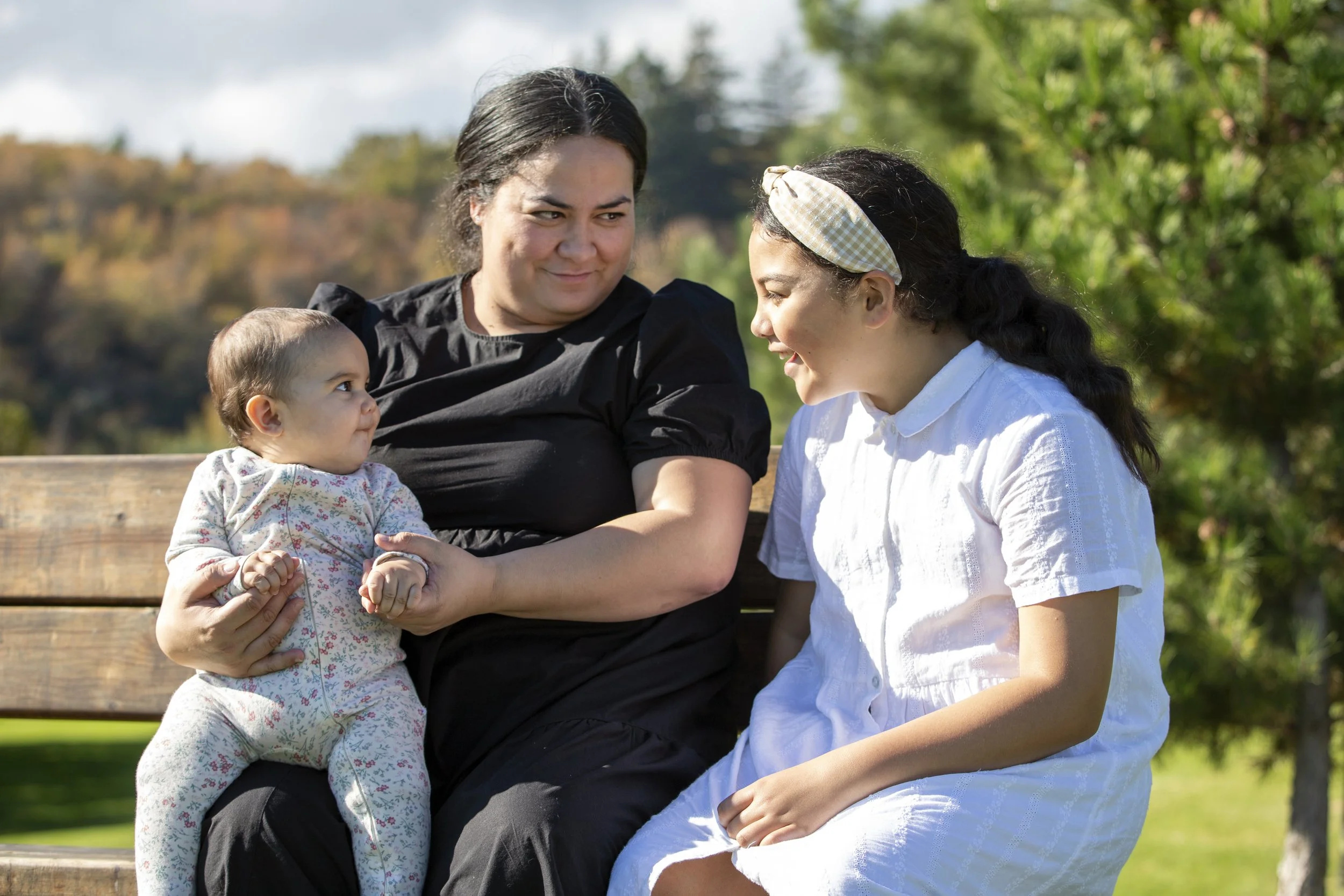 Three women sitting on a park bench, engaging and smiling at each other, with trees and sky in the background.