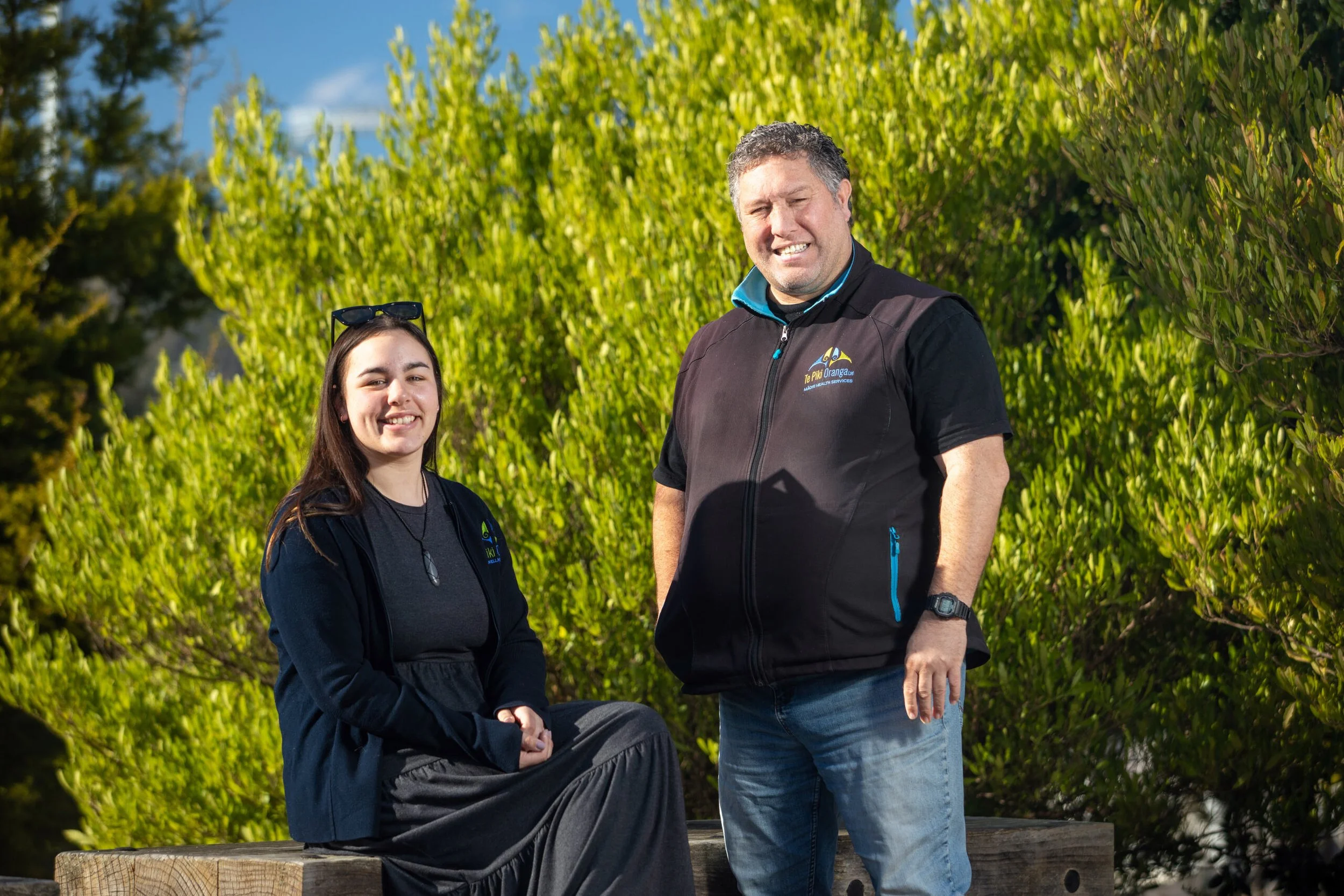 A young woman sitting on a wooden bench, smiling, with an older man standing beside her outdoors with lush green bushes and trees in the background.