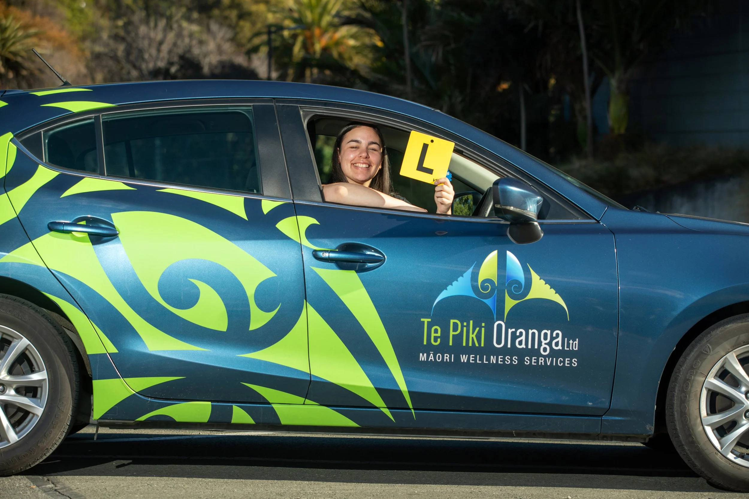 A woman sitting in a blue car with green decorative patterns, holding a yellow learner's permit sign and smiling. The car has the logo and text 'Te Piki Oranga Ltd Māori Wellness Services' on the side.