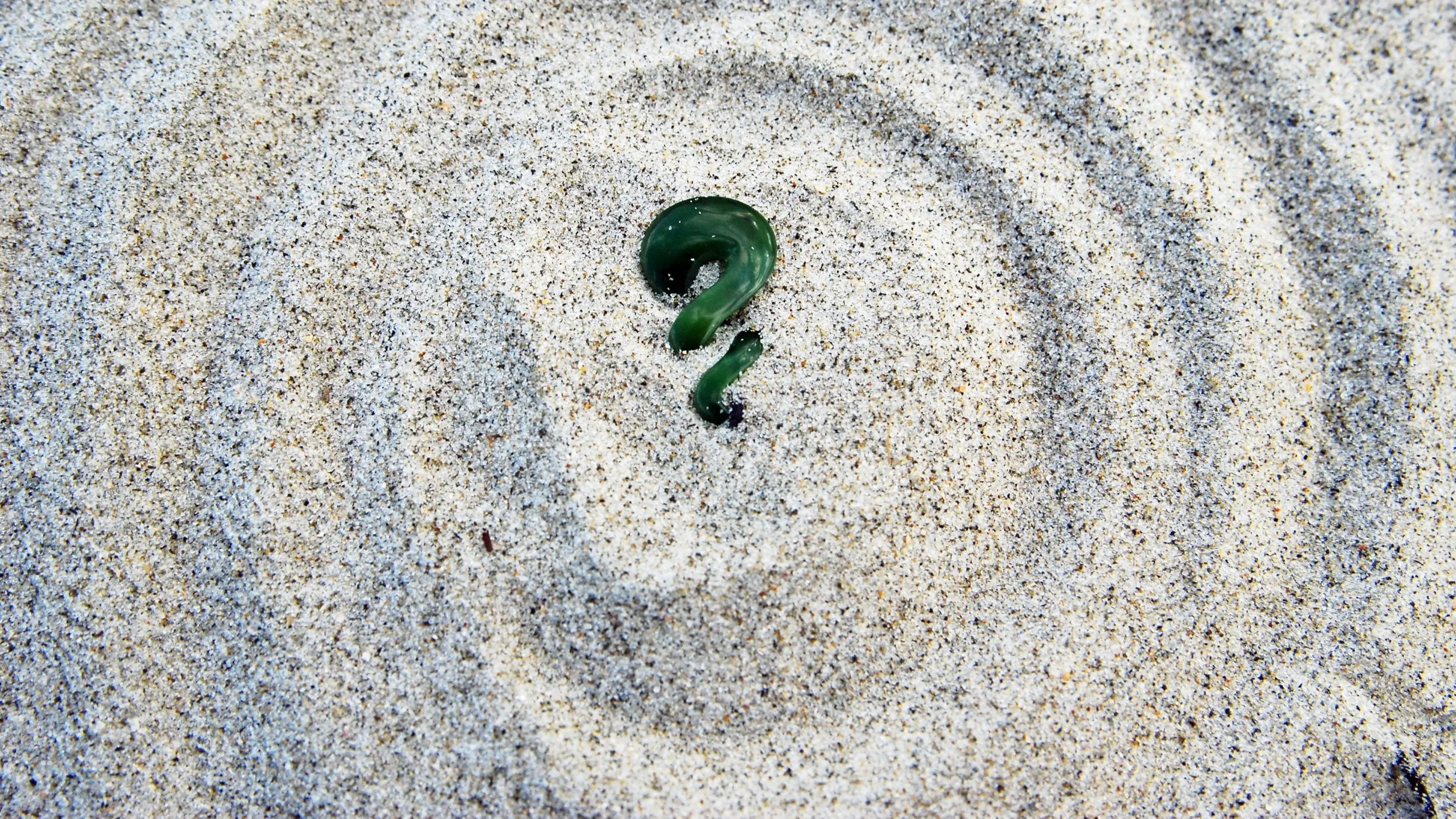 A green question mark-shaped object partially buried in light-colored sand, with visible shadow.