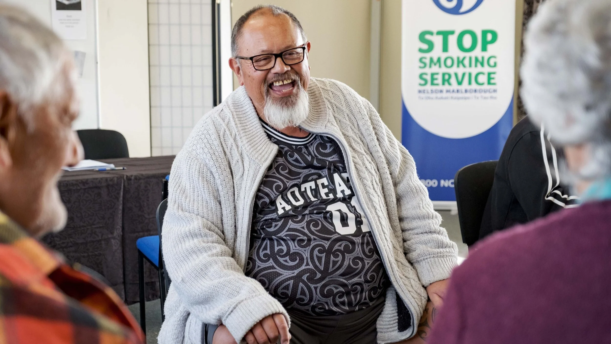 A smiling man with glasses and a beard, wearing a gray sweater and a sports jersey, sitting at a table with other elderly people in a community center, with a sign in the background that reads 'Stop Smoking Service'.