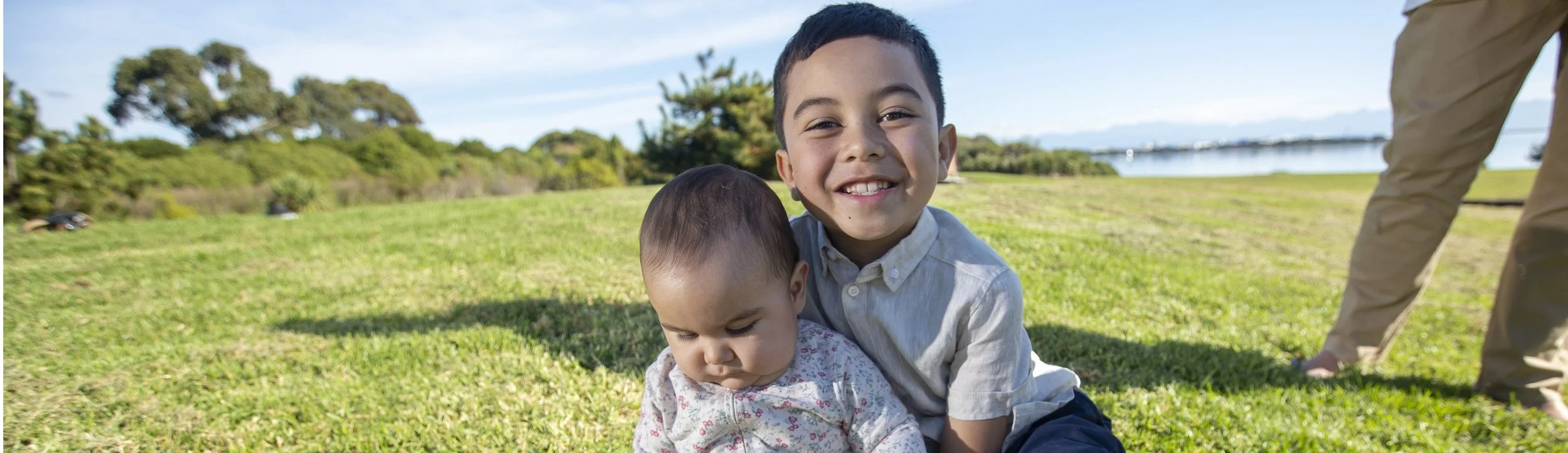 Two young boys, one older and one younger, sitting on grass in a park with trees and water in the background, during daytime.