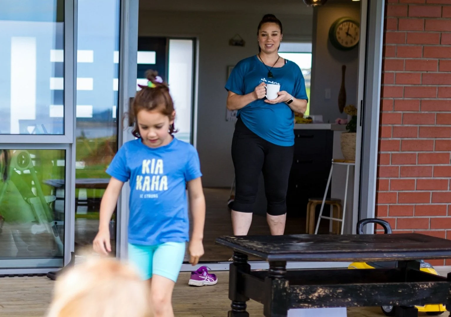 A young girl wearing a blue T-shirt and light blue shorts walking on a wooden deck, with a woman in the background holding a white mug and standing inside a house near an open sliding glass door.