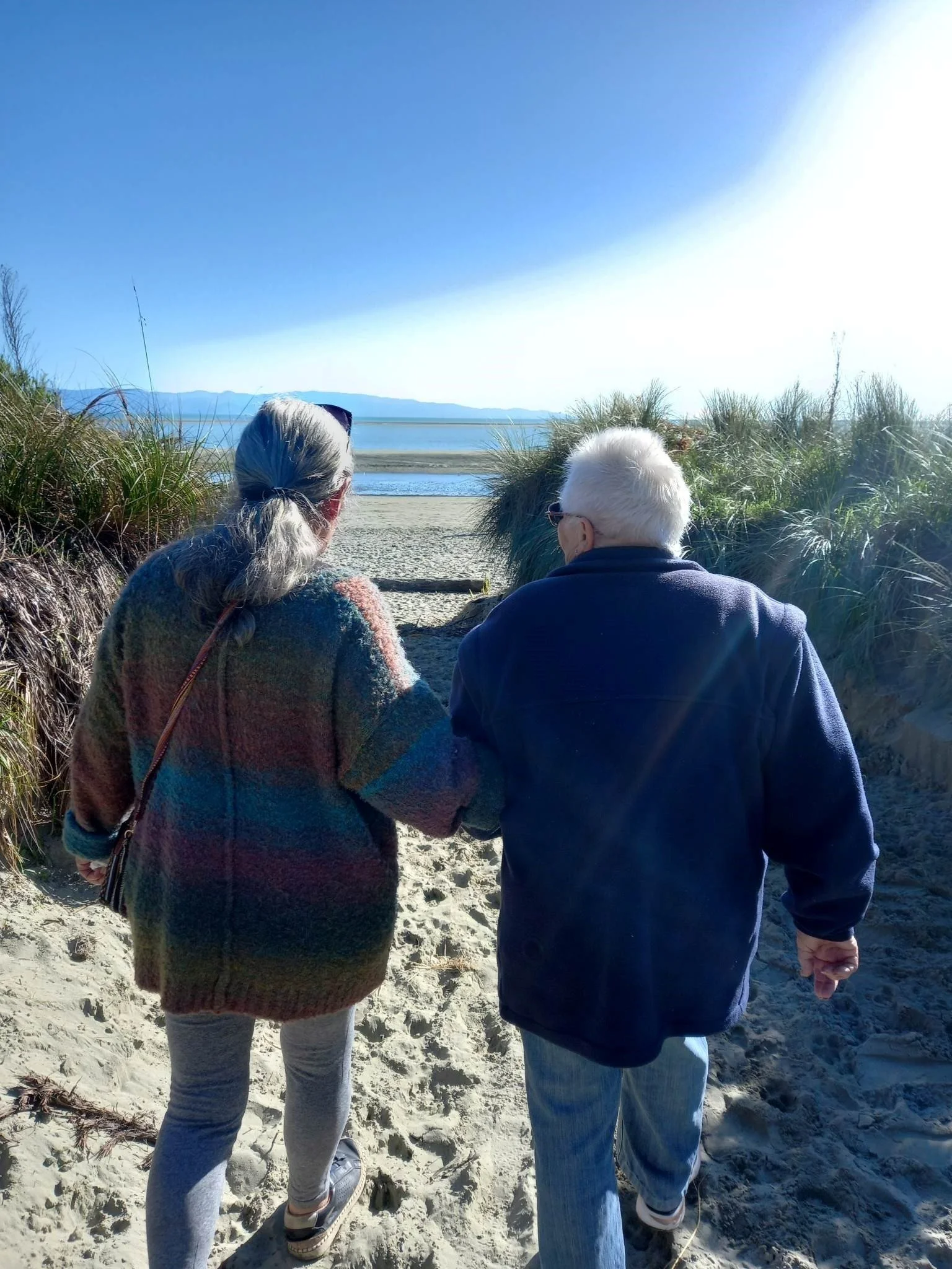Older woman and woman walking hand-in-hand towards a beach with water and mountains in the distance, surrounded by sand dunes and tall grass under a clear blue sky.
