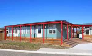 A single-story building with large windows, red framing, and a wooden ramp leading to the entrance, under a blue sky.