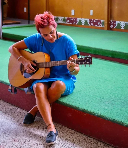 A woman with short pink hair sitting cross-legged on a stage, playing an acoustic guitar.