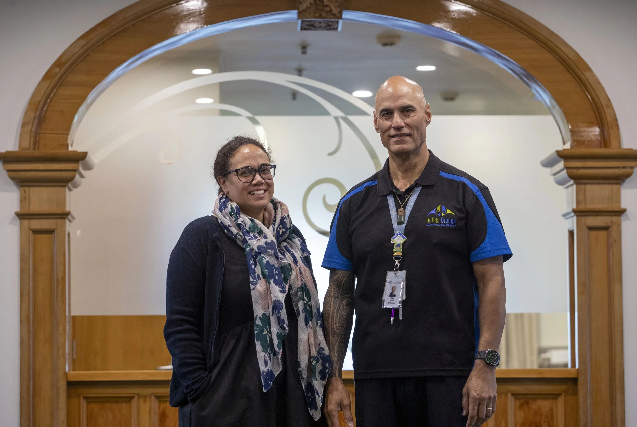 Two kaimahi (staff) stand beneath a wooden archway at Te Piki Oranga whare on Nayland Rd.