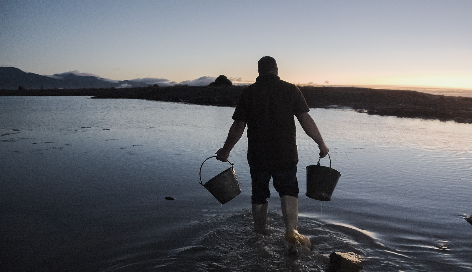A woman and a man with traditional tattoos on their faces and necks standing outdoors near water, smiling at the camera during daytime.