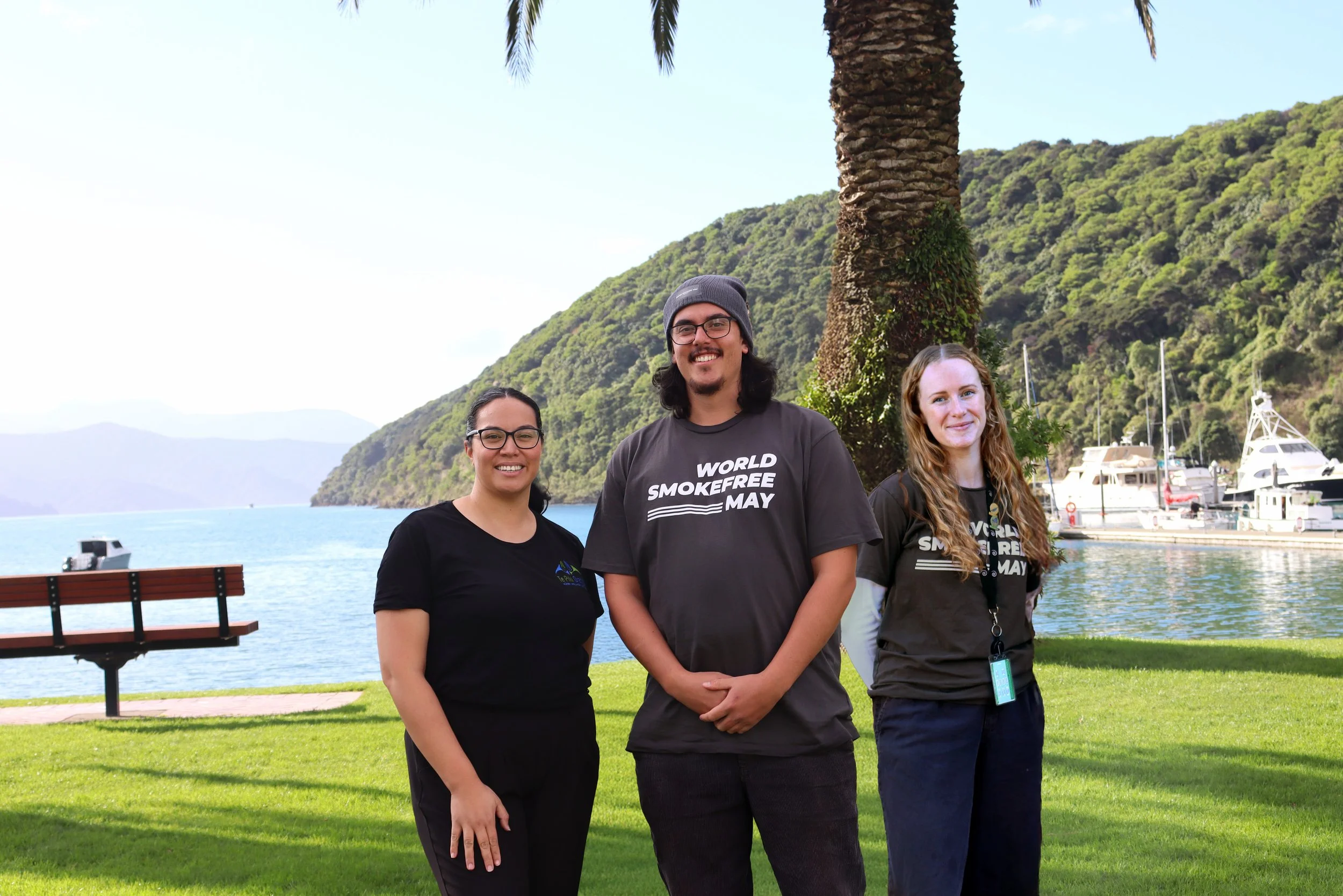 Three people standing in front of a large palm tree and a body of water with boats, mountains, and a clear sky in the background. All are smiling, with the two women on either side wearing black shirts, and the man in the middle wearing a dark gray t-shirt with the text "WORLD SMOKEFREE MAY."