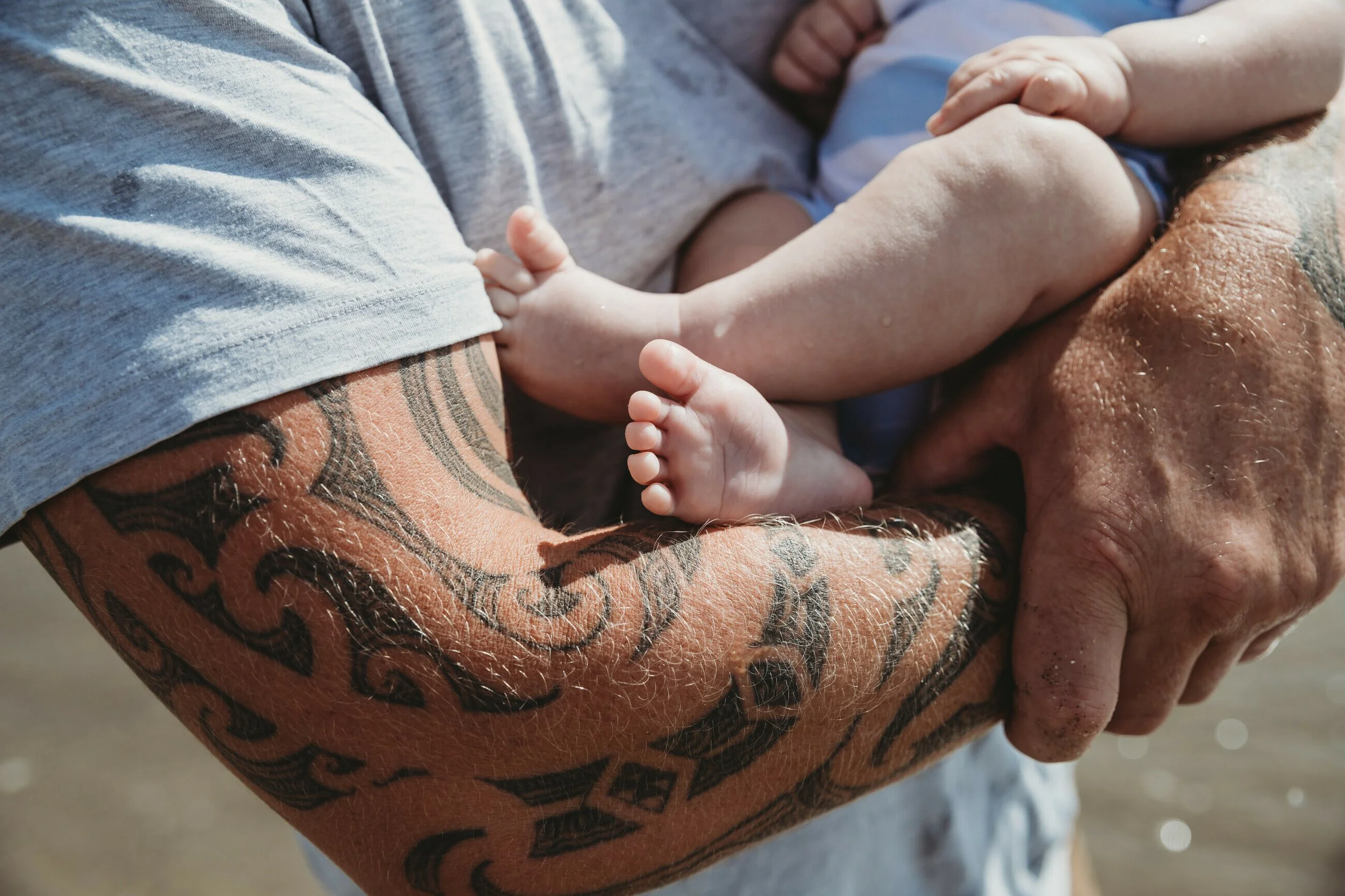 A person's tattooed arm holding a small child's arm and hand, with the child sitting on the person's lap, outdoors on a sunny day.