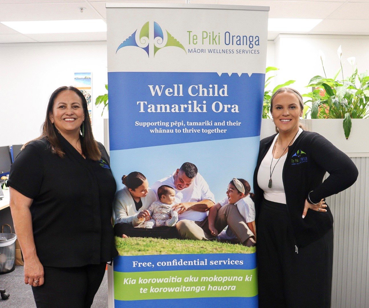 Two smiling wahine (women) from Te Piki Oranga stand beside a promotional banner for “Well Child Tamariki Ora”.